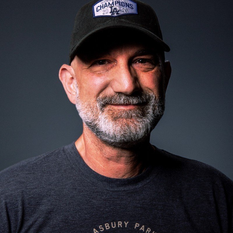 A man with a beard and mustache wearing a black baseball cap and a dark T-shirt, smiling.