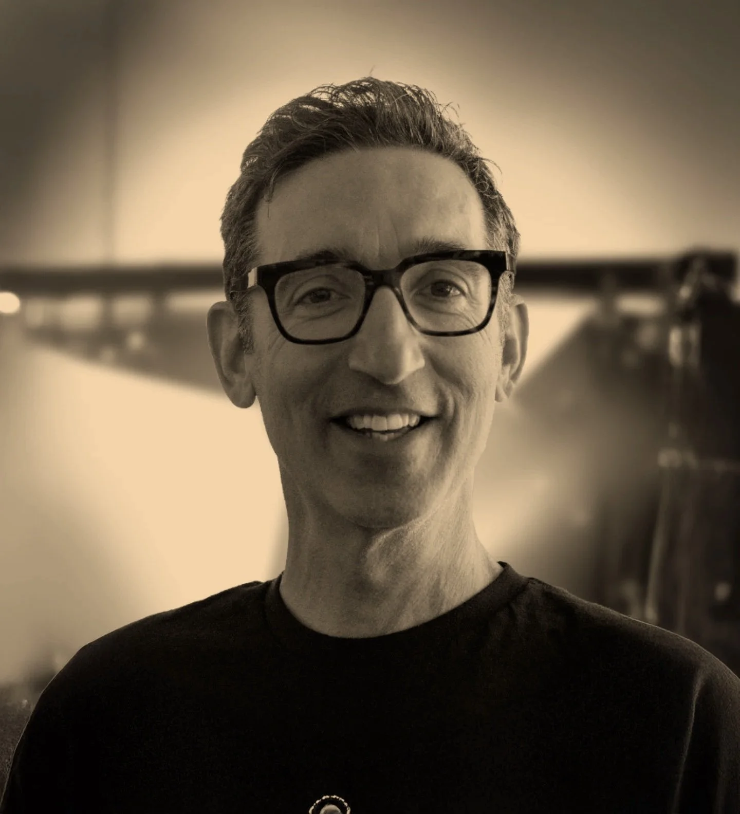 A black-and-white photo of a smiling man with glasses, short dark hair, wearing a black shirt, standing indoors.