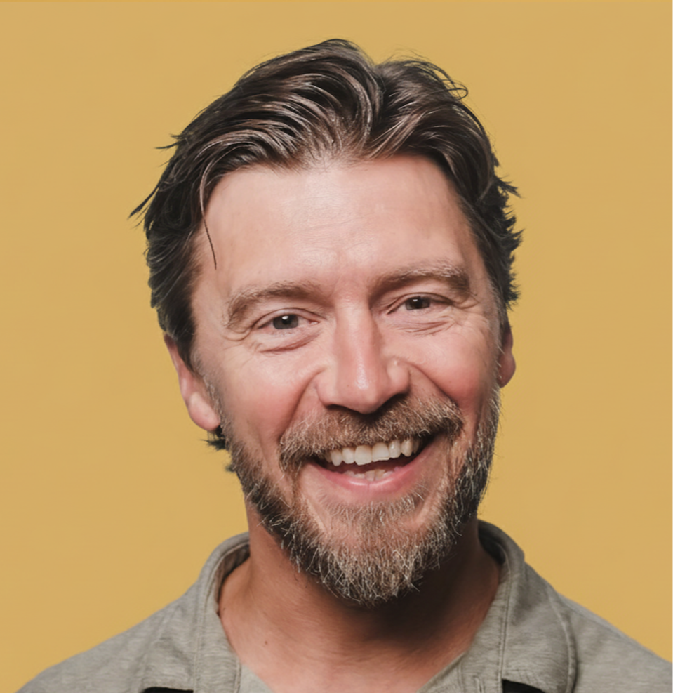 Close-up of a middle-aged man with brown hair, a beard, smiling, wearing a gray shirt, against a yellow background.