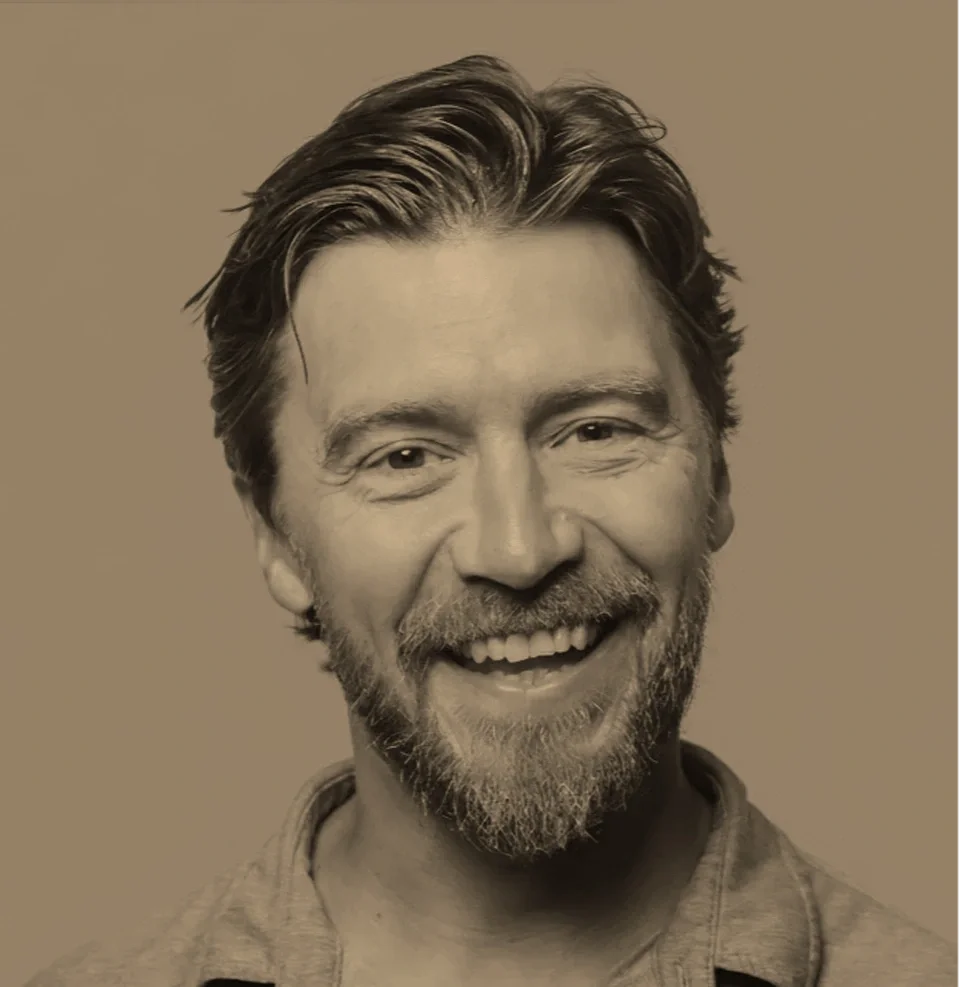 A black and white portrait of a smiling man with a beard and wavy hair, wearing a casual shirt.