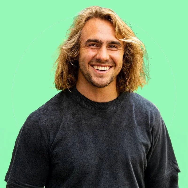 A smiling man with long blonde hair wearing a black T-shirt against a light green background.