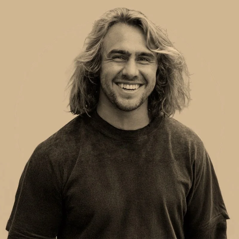 A young man with long, wavy hair and a beard, smiling at the camera, wearing a dark t-shirt against a plain background.