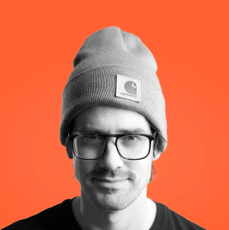 A black-and-white portrait of a man wearing glasses and a beanie hat, set against an orange background.
