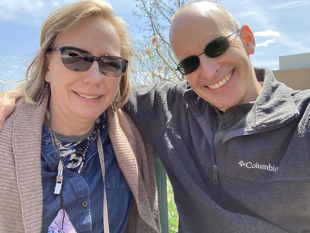 A woman and a man smiling outdoors, both wearing sunglasses, with the woman having shoulder-length blonde hair and the man being bald with a broad smile. The woman is dressed in a blue denim shirt and a light brown cardigan, while the man is in a black Columbia jacket. The background shows a clear blue sky and some trees.