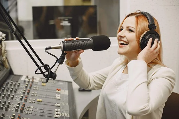 A woman with red hair wearing headphones, speaking into a microphone, in a radio studio with a mixing console.