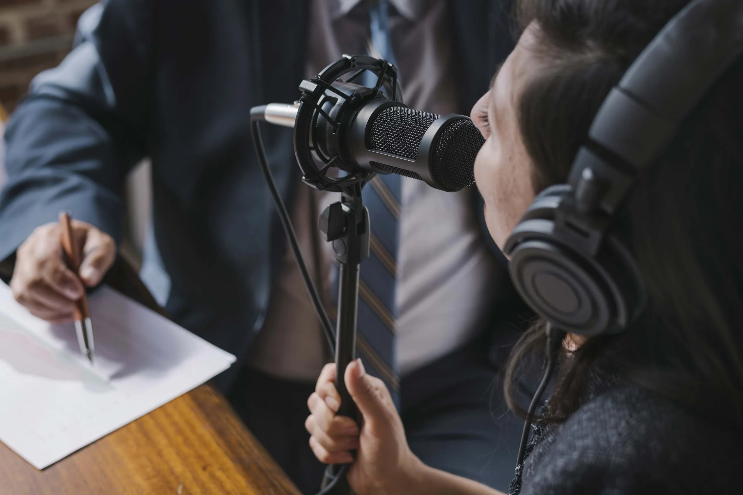 Close-up of a woman with headphones speaking into a microphone while a person in formal attire writes on a paper.