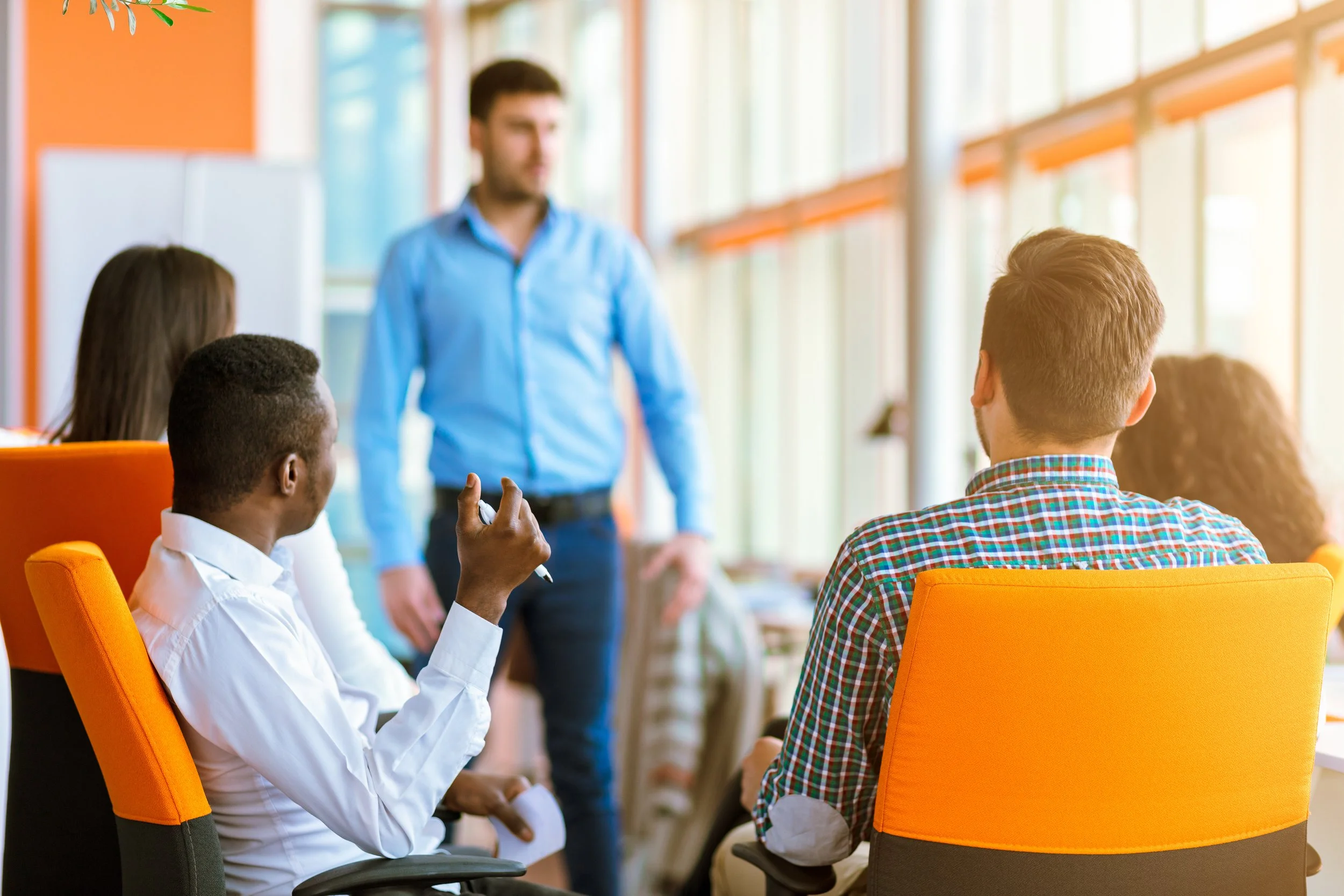A group of diverse people having a discussion in a bright, modern office or conference room. The focus is on a man in a white shirt holding a pen, facing an individual in a plaid shirt. In the background, a man in a blue shirt appears to be speaking or presenting.