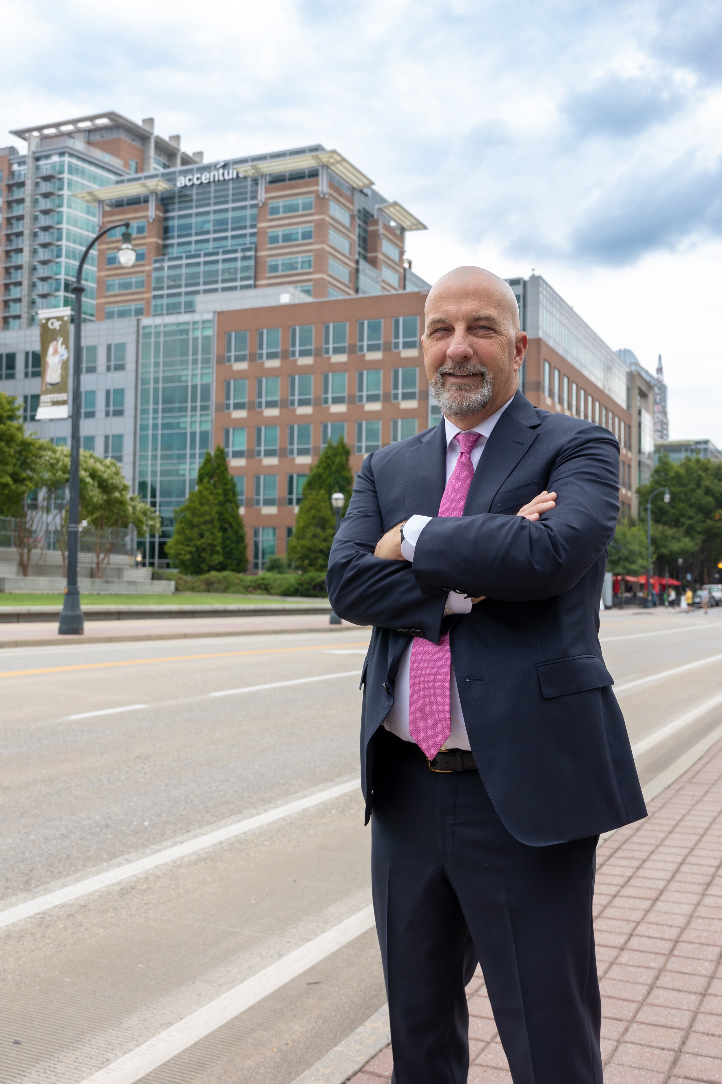 A man in a suit with a pink tie standing on a city sidewalk with arms crossed, office buildings, trees, and a cloudy sky in the background.