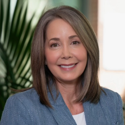 A smiling woman with shoulder-length brown hair and light skin, wearing a blue blazer, standing indoors with a large green plant and a bright window in the background.