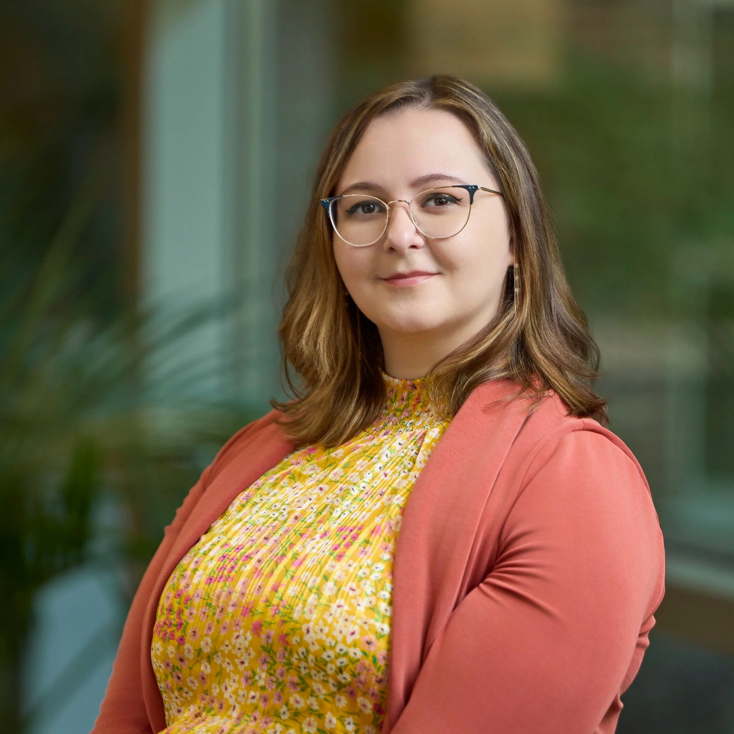 A woman with shoulder-length brown hair and glasses, wearing a yellow floral dress and a pink blazer, posing indoors near a window with greenery in the background.
