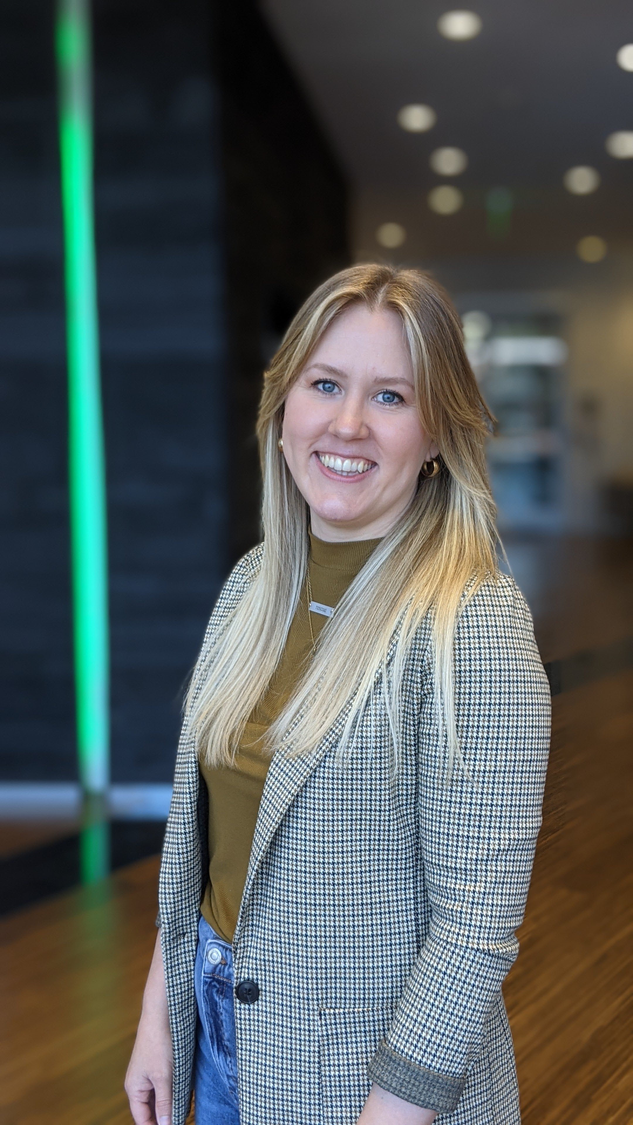 A woman with long blonde hair, blue eyes, and a bright smile standing indoors.