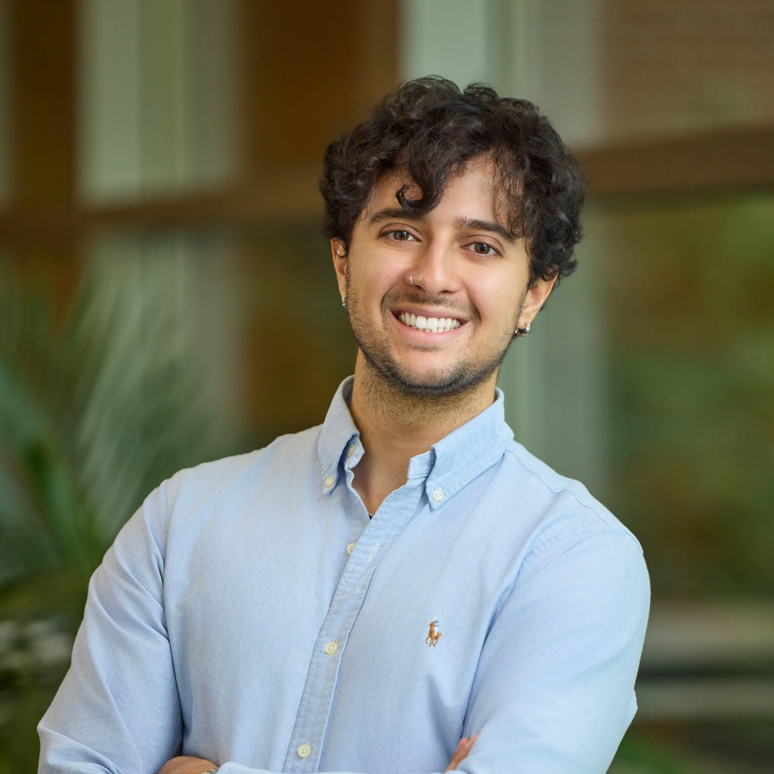 A young man with curly dark hair, earrings, a nose ring, and a light blue button-up shirt, smiling with arms crossed in an indoor setting.