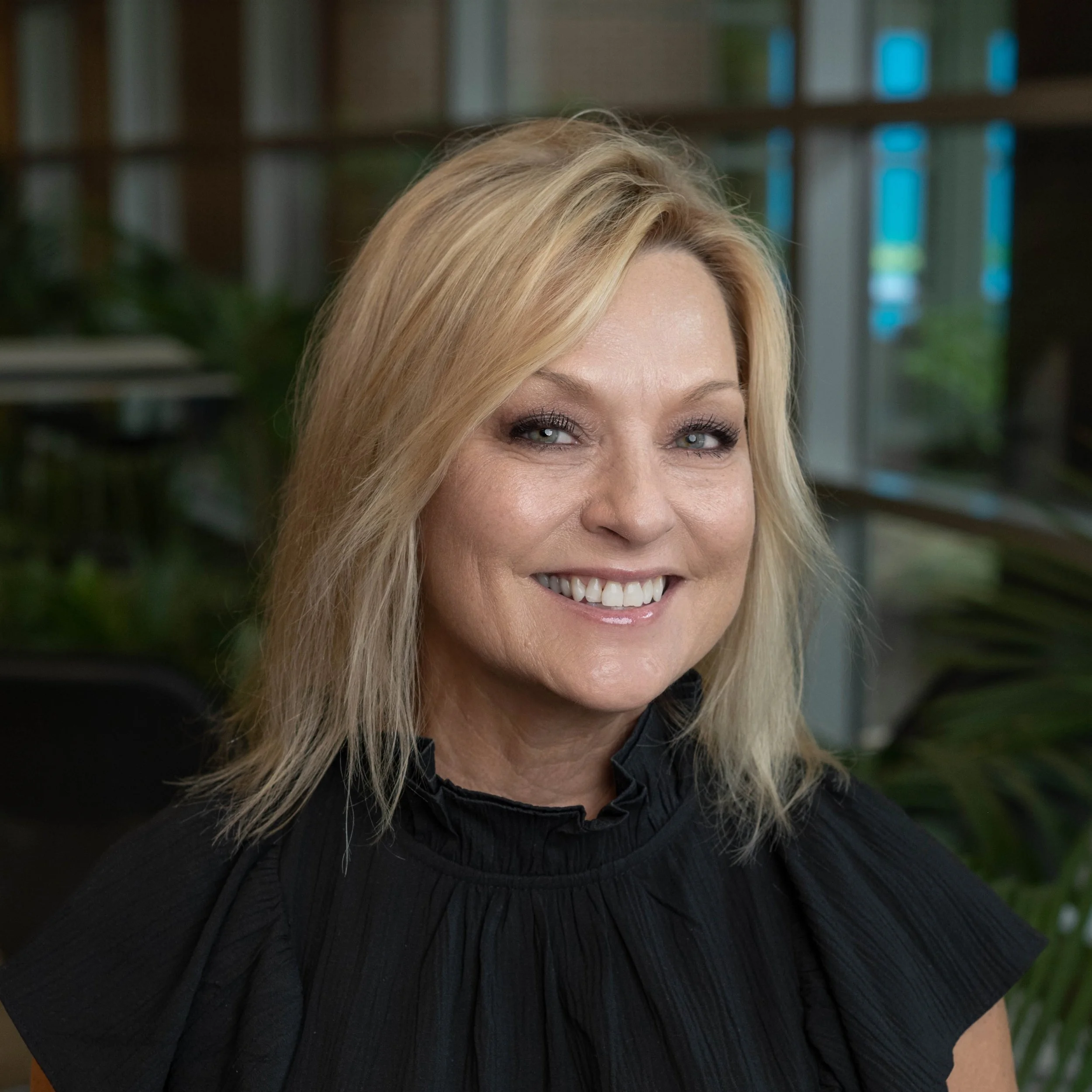 Portrait of a smiling blonde woman with blue eyes, wearing a black ruffled top, indoors with greenery and large windows in the background.