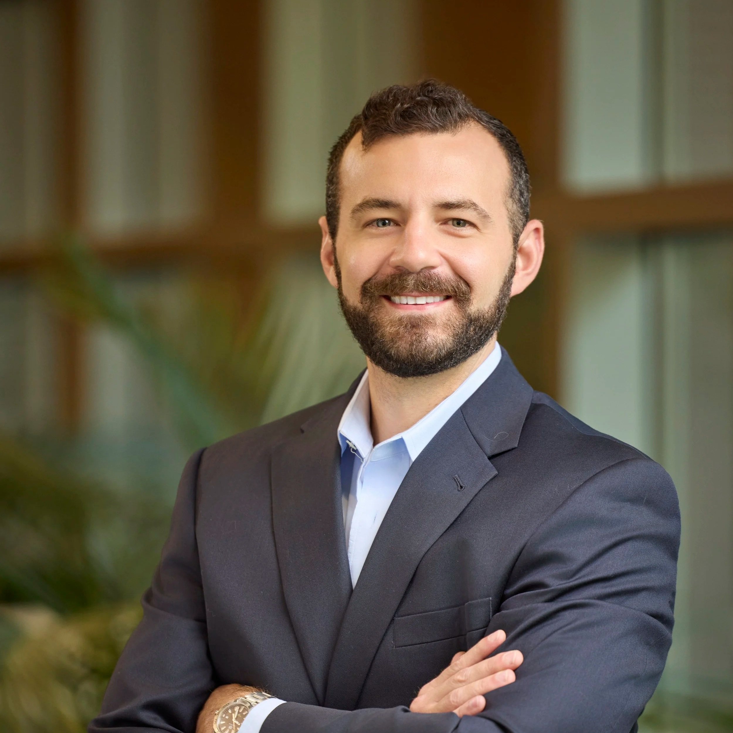 Professional man smiling, wearing a dark suit and white shirt, arms crossed, indoor setting with blurred shelves or windows in the background.