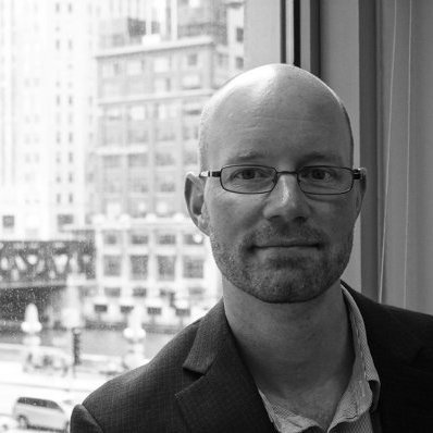 A black and white photo of a man with glasses, a beard, and a bald head, sitting indoors near a window with a cityscape in the background.