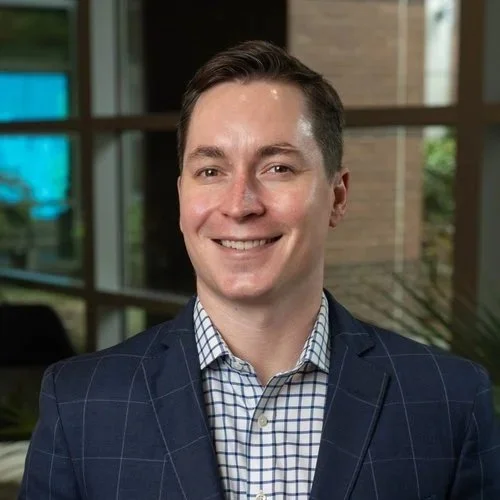 Portrait of a smiling man in a business suit inside a modern office building with large windows.