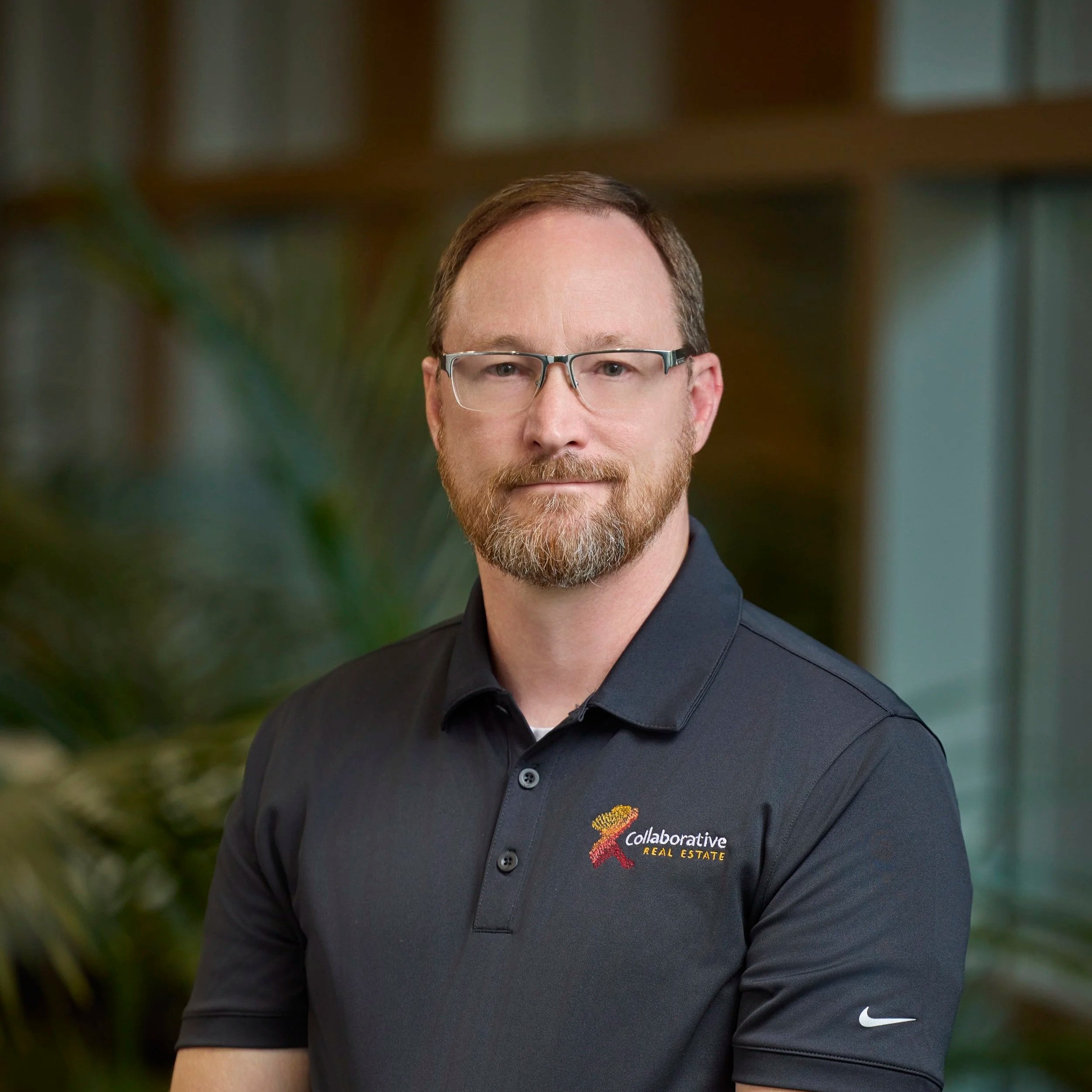 A man with glasses, a beard, and short hair wearing a black polo shirt with the logo of Collaborative Real Estate, standing indoors with a blurred background of wooden shelving and a plant.
