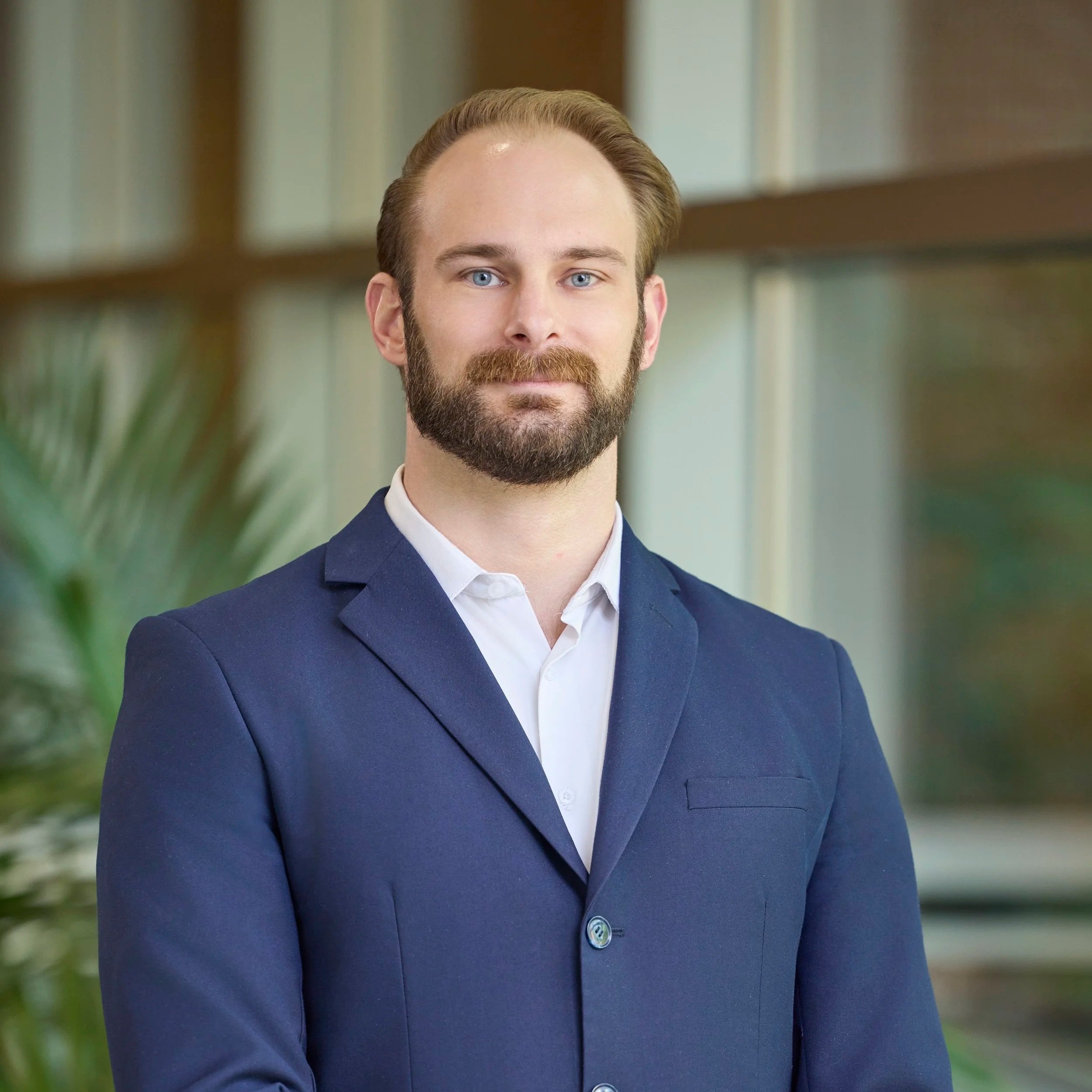 A man in a navy blue suit with a white shirt standing in an indoor setting with blurred background and green plant on the side.