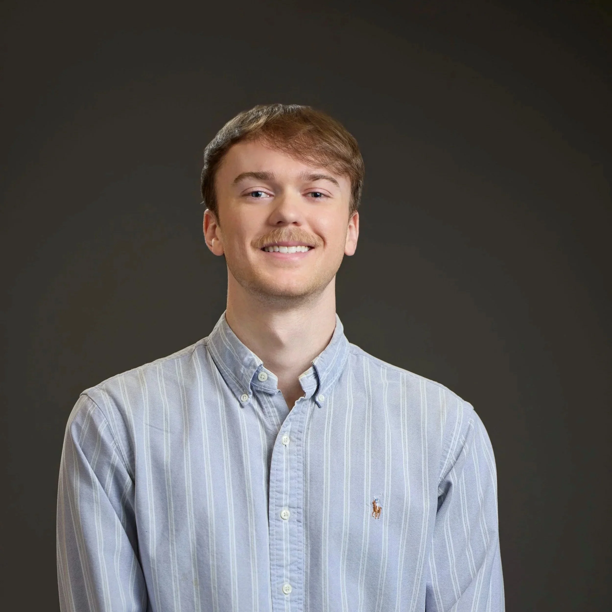 Portrait of a young man with light brown hair, a mustache, and blue eyes, smiling, wearing a light blue striped button-up shirt with a Ralph Lauren logo, against a dark background.