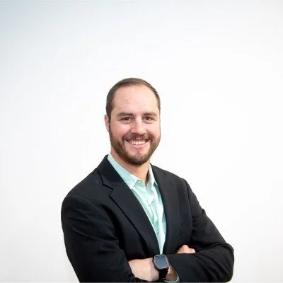 A smiling man with a beard, wearing a black blazer and light blue shirt, standing against a white background.
