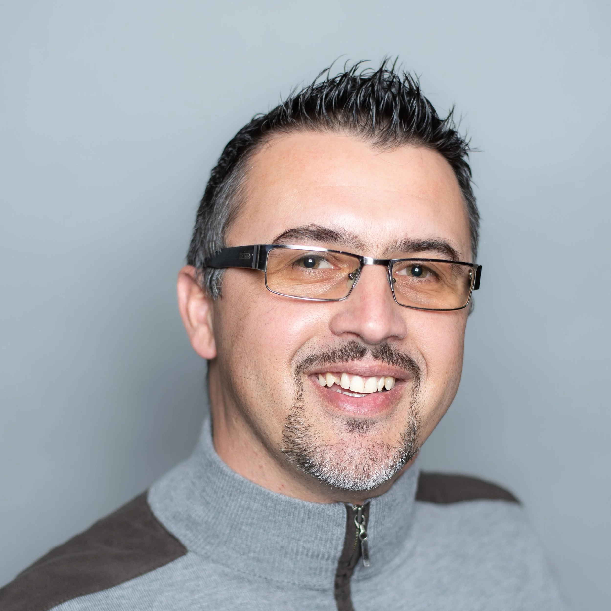 A middle-aged man with short, dark hair, glasses, and facial hair, smiling at the camera against a plain background.