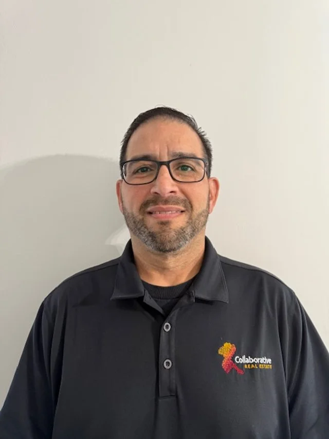 Man with glasses and short dark hair, wearing a collared black shirt with a logo on the chest, standing against a plain white wall.