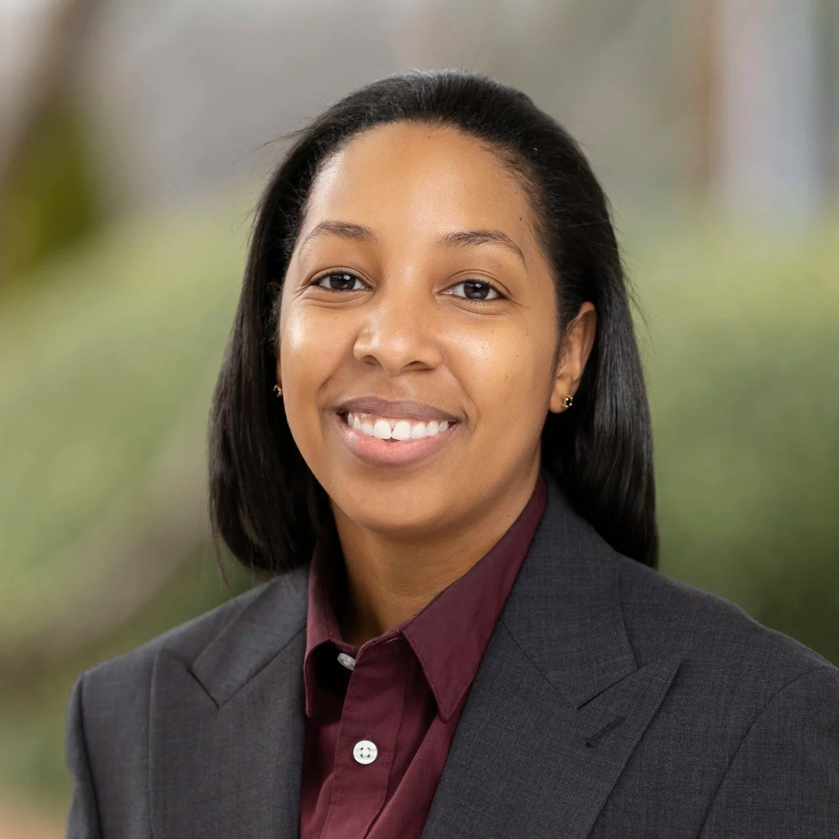 A smiling woman with dark hair, wearing a gray blazer and maroon blouse, posing outdoors.