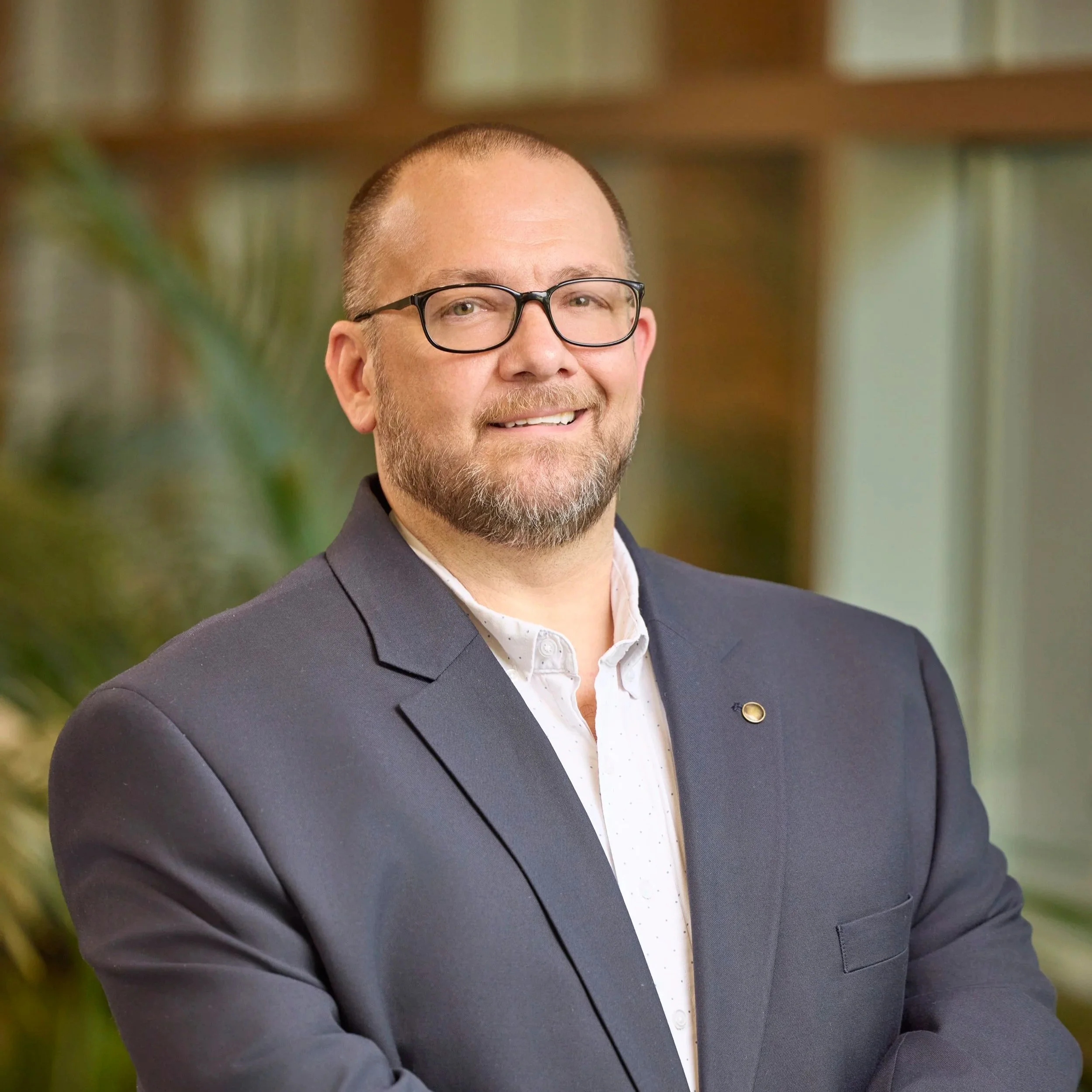 Portrait of a man with glasses, short hair, and beard, dressed in a dark suit jacket and white shirt, smiling indoors with a blurred background of greenery and wooden shelves.