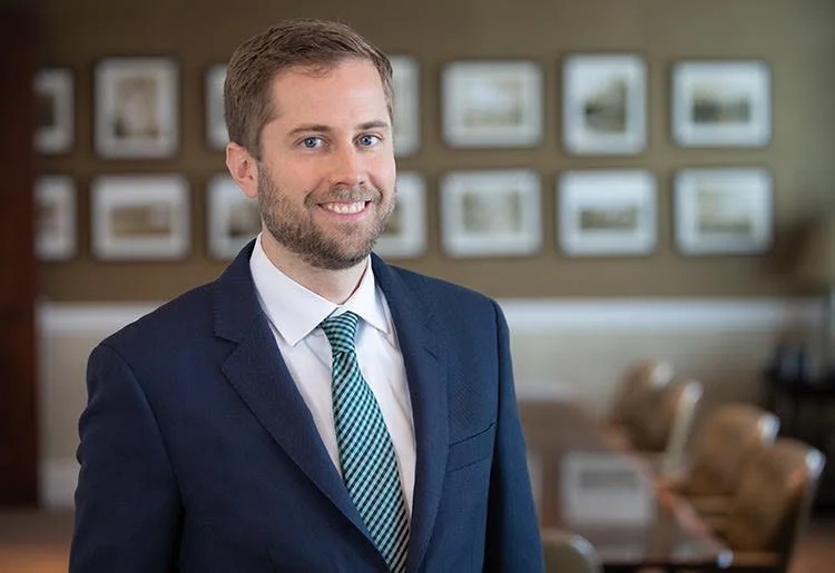 A man with a beard and light brown hair smiling, dressed in a navy suit, white shirt, and a teal striped tie, standing in an office or conference room with framed pictures on the wall and brown chairs in the background.