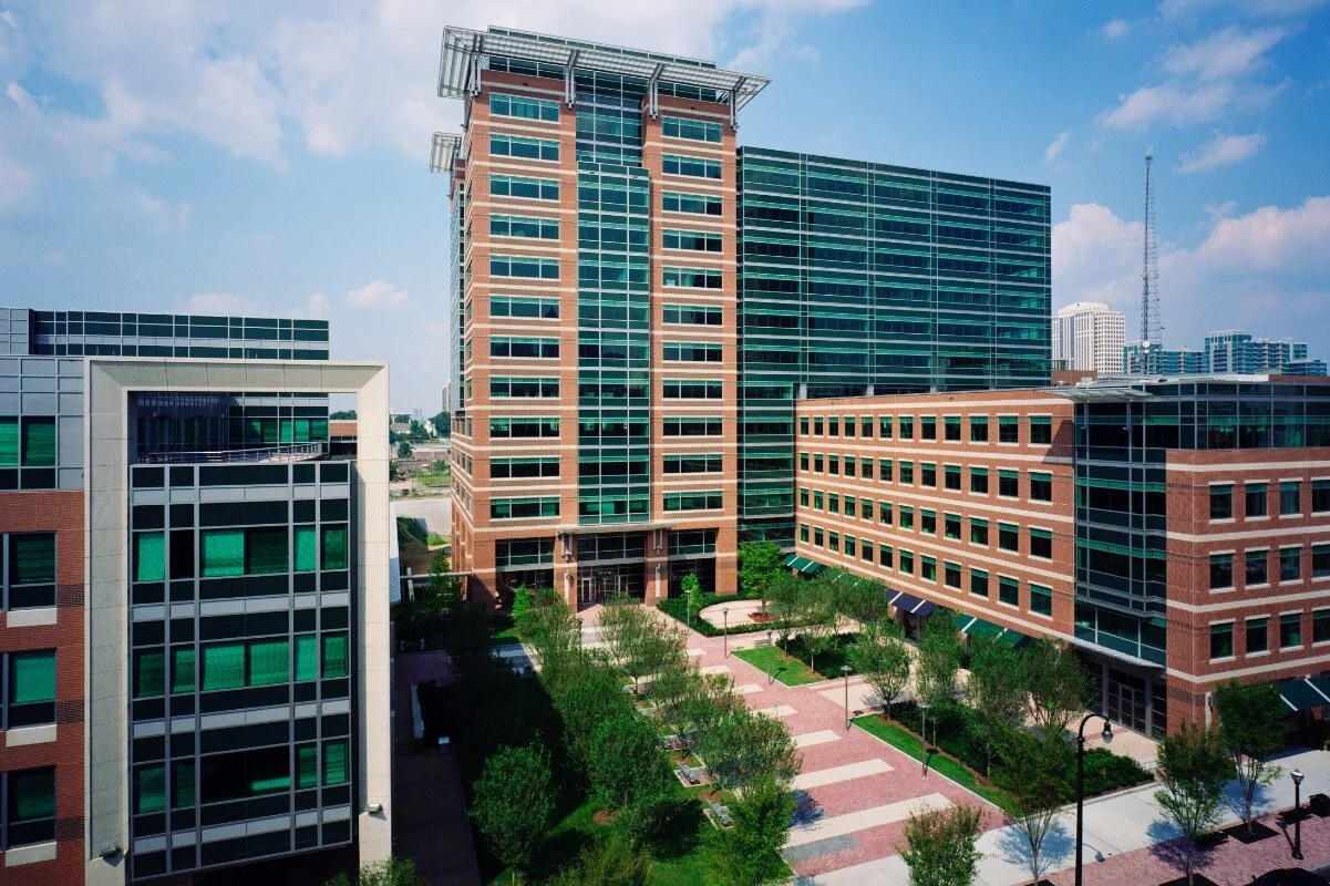 High-rise office buildings with glass windows in an urban area, greenery, and a paved walkway in the foreground.