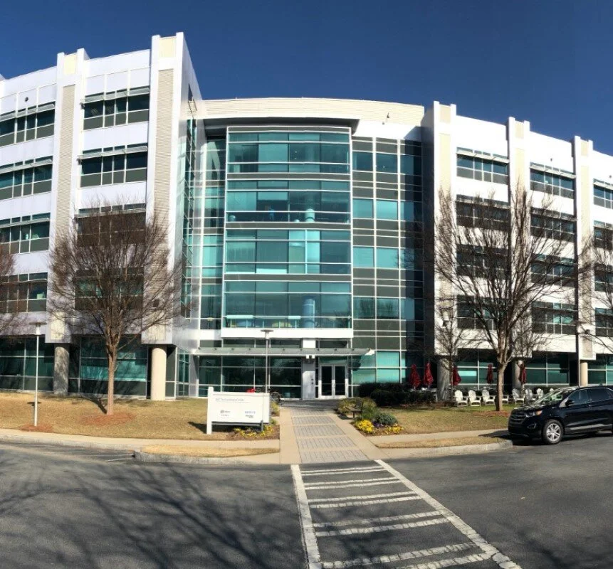 Modern multi-story office or apartment building with glass windows, trees, and parked cars in front.