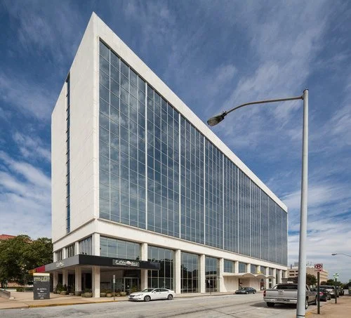 A tall modern office building with large glass windows reflecting the sky, situated on a city street with cars parked in front.