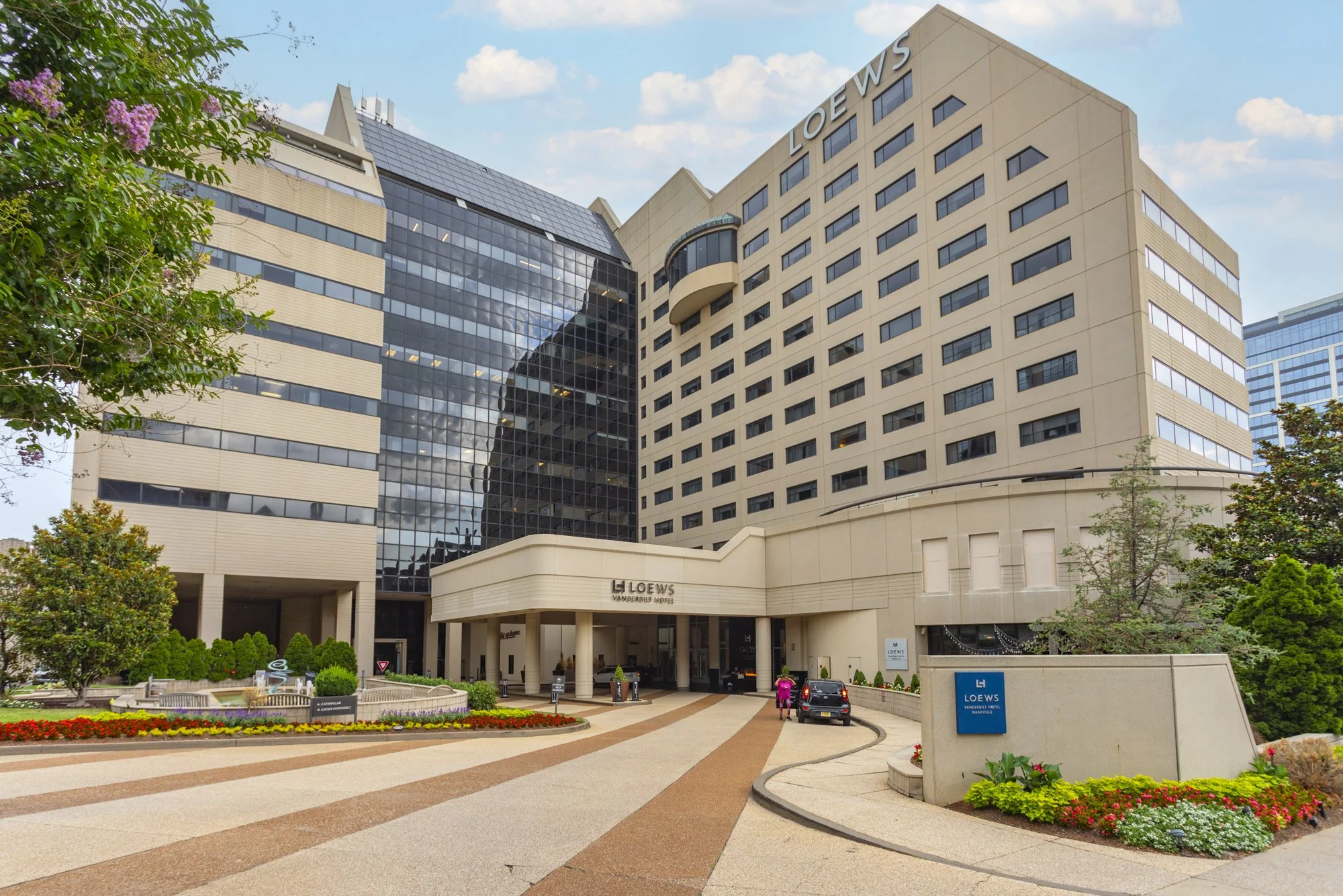 Exterior view of the Loews Vanderbilt Hotel in Nashville, showing a modern multi-story building with a mix of beige and glass facade, surrounded by greenery and a landscaped area in the front, with a driveway leading to the entrance.