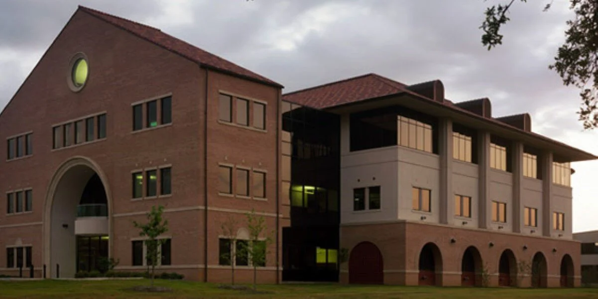 A multi-story building with a brick facade, arched entryway, and large windows, set outdoors with small trees and cloudy sky.
