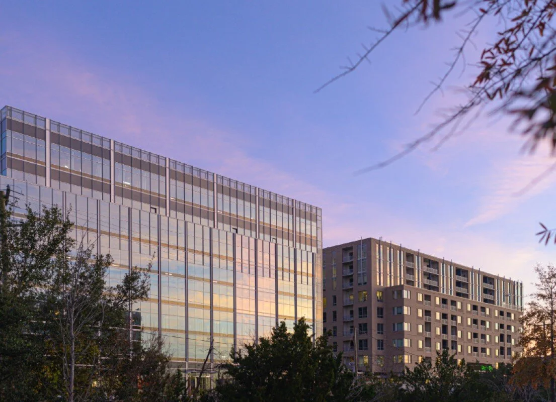 Glass office building reflecting sunset with surrounding trees and a neighboring residential building.