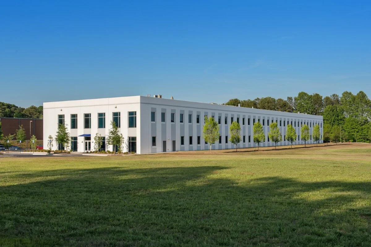 A modern white office building with multiple windows, surrounded by a grassy lawn and young trees, under a clear blue sky.