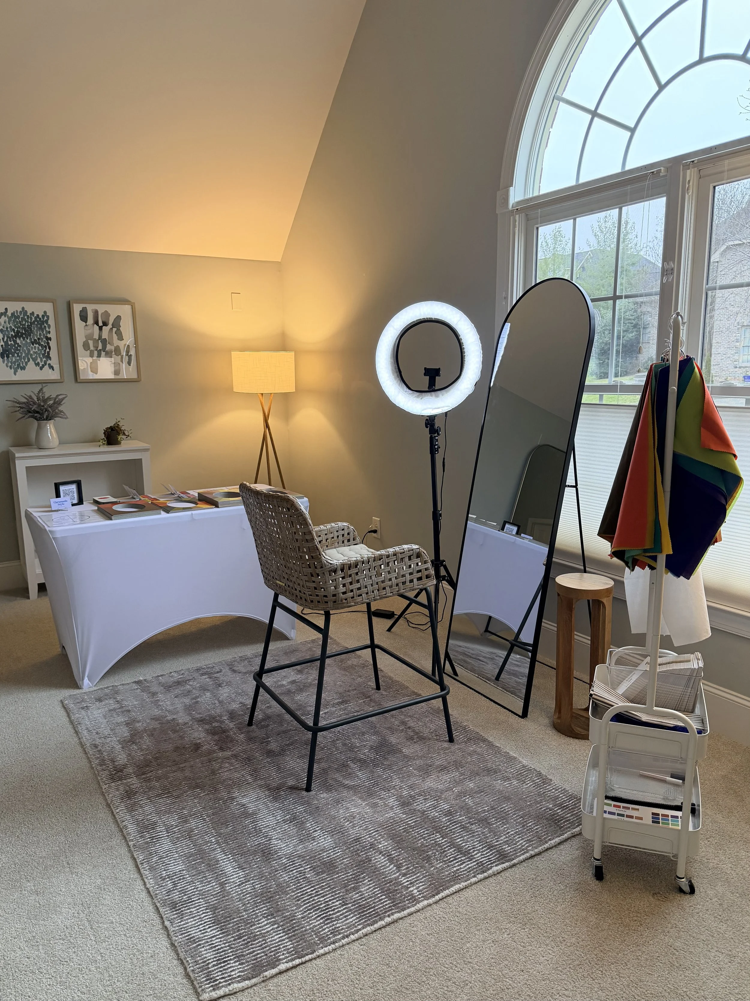 A room setup with a large arched window, a ring light, a mirror, a wicker chair, and a table with various color analysis frames, including colorful drapes hanging on a stand.