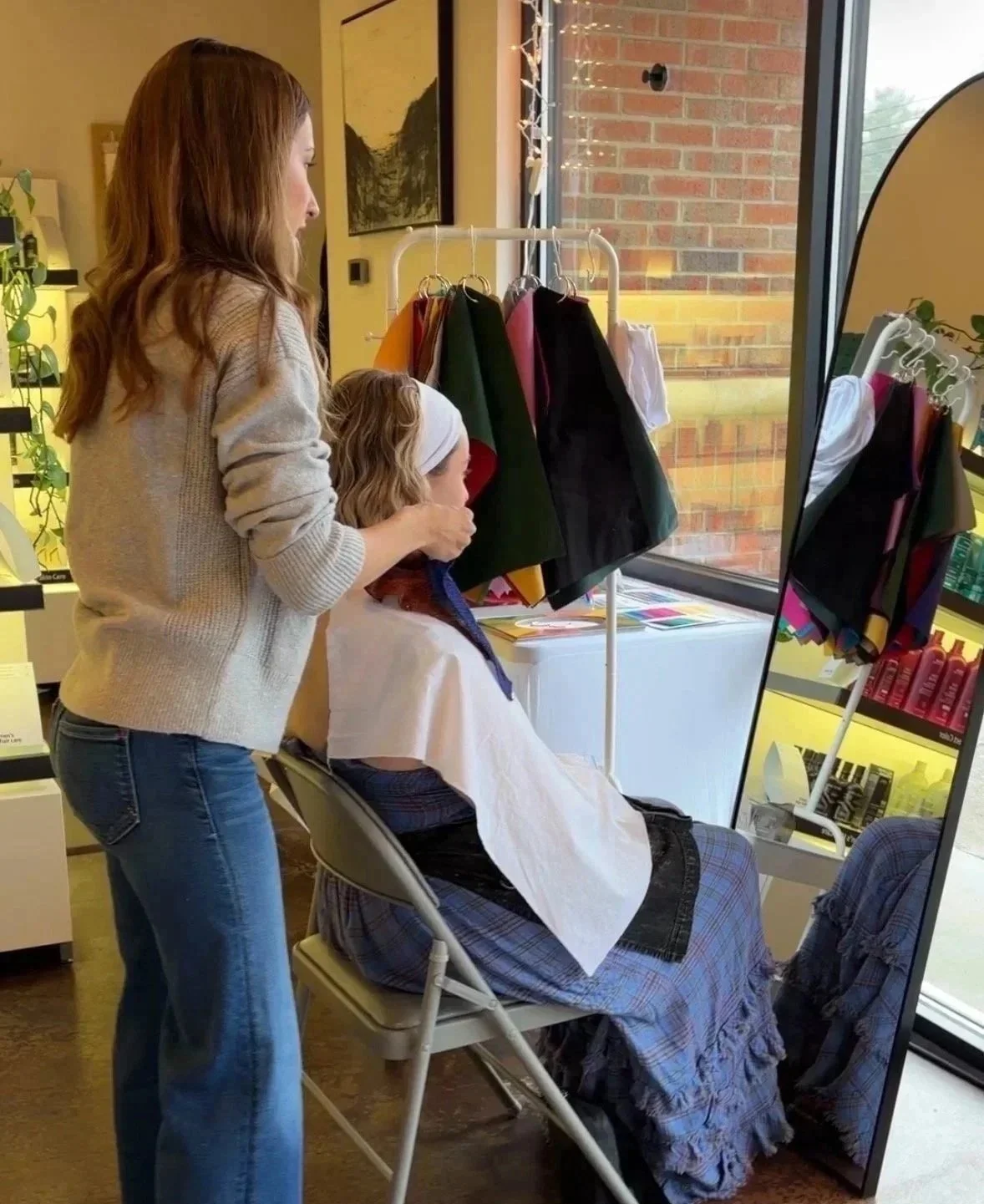 Woman getting her seasonal color analysis done styled by a female color analyst in a salon, sitting in front of a large mirror, with color analysis color drapes over her shoulders.