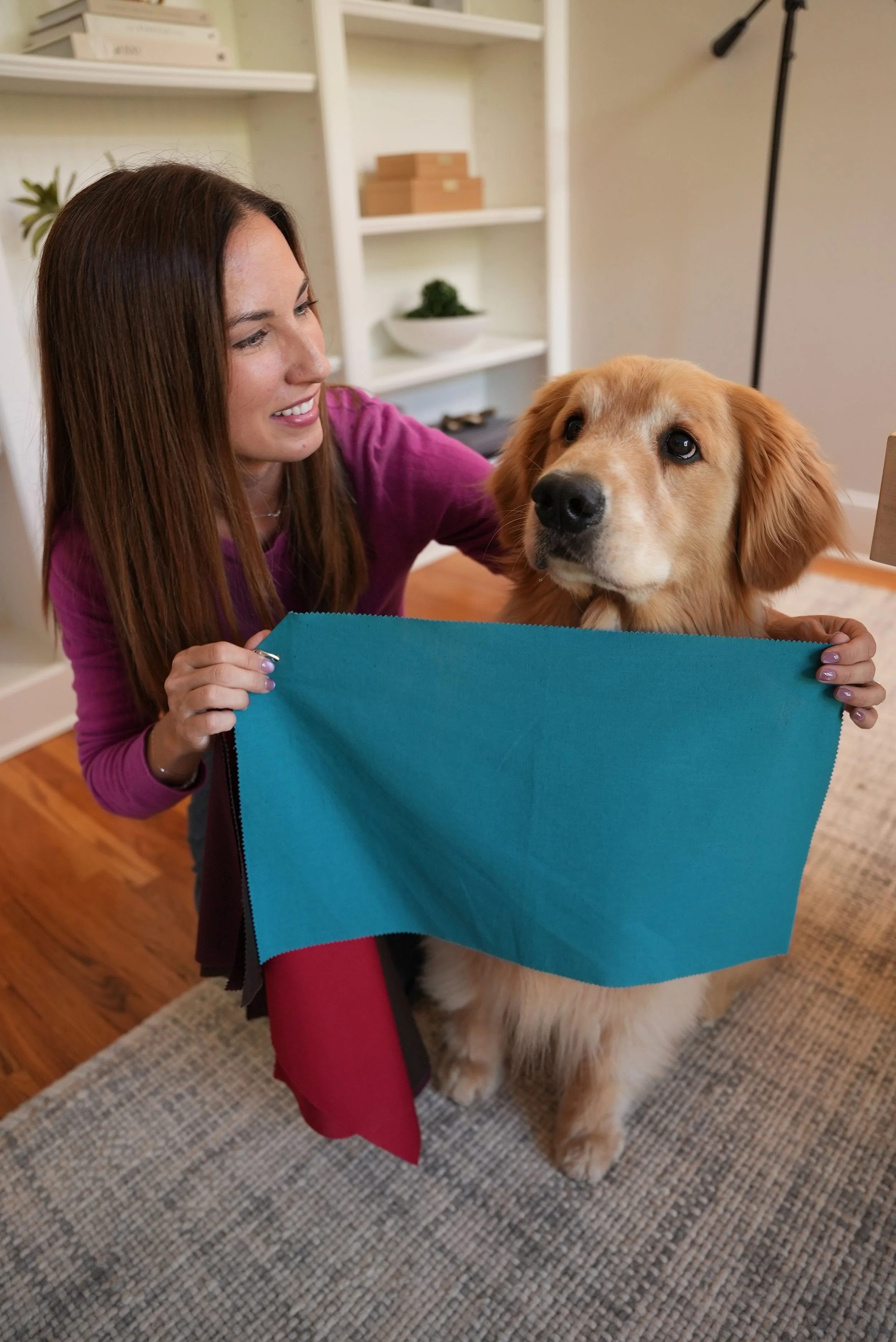 A woman with long brown hair, wearing a pink top, holding a blue fabric color analysis drape in front of a golden retriever indoors.