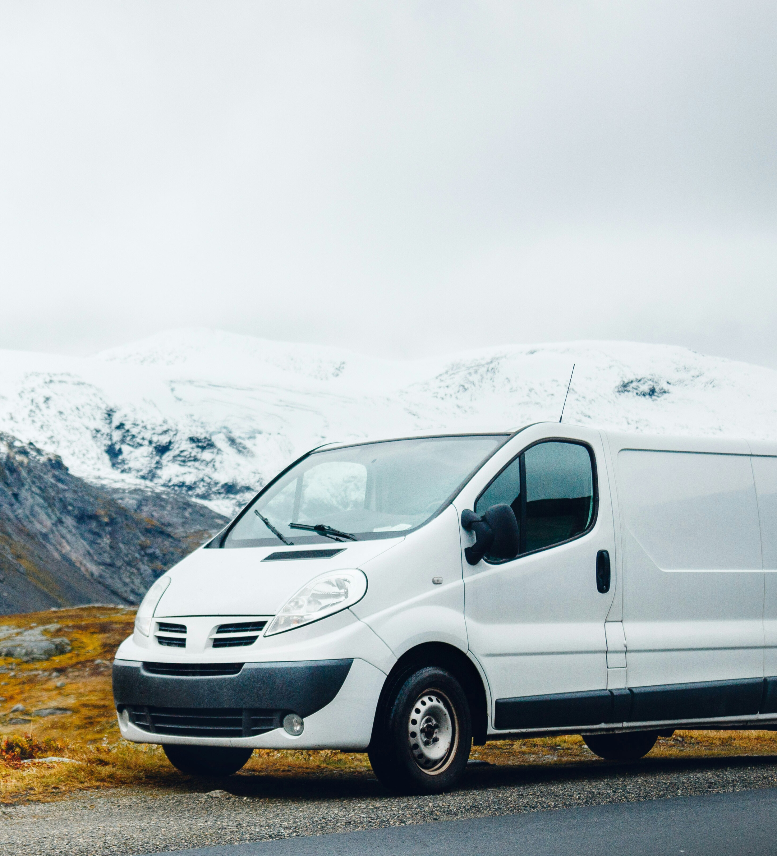 A white cargo van parked on the side of a mountain road with snow-capped mountains in the background.