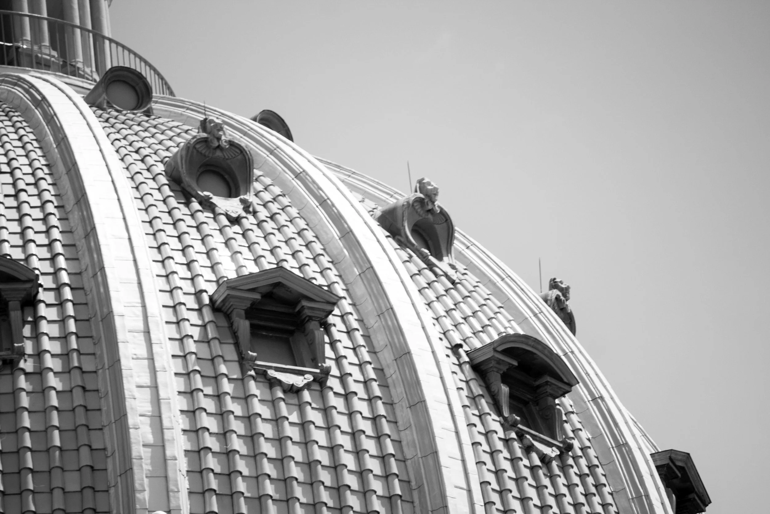 Close-up of a decorative, tiled dome roof with ornate architectural details and small windows, against a plain sky background, in black and white.