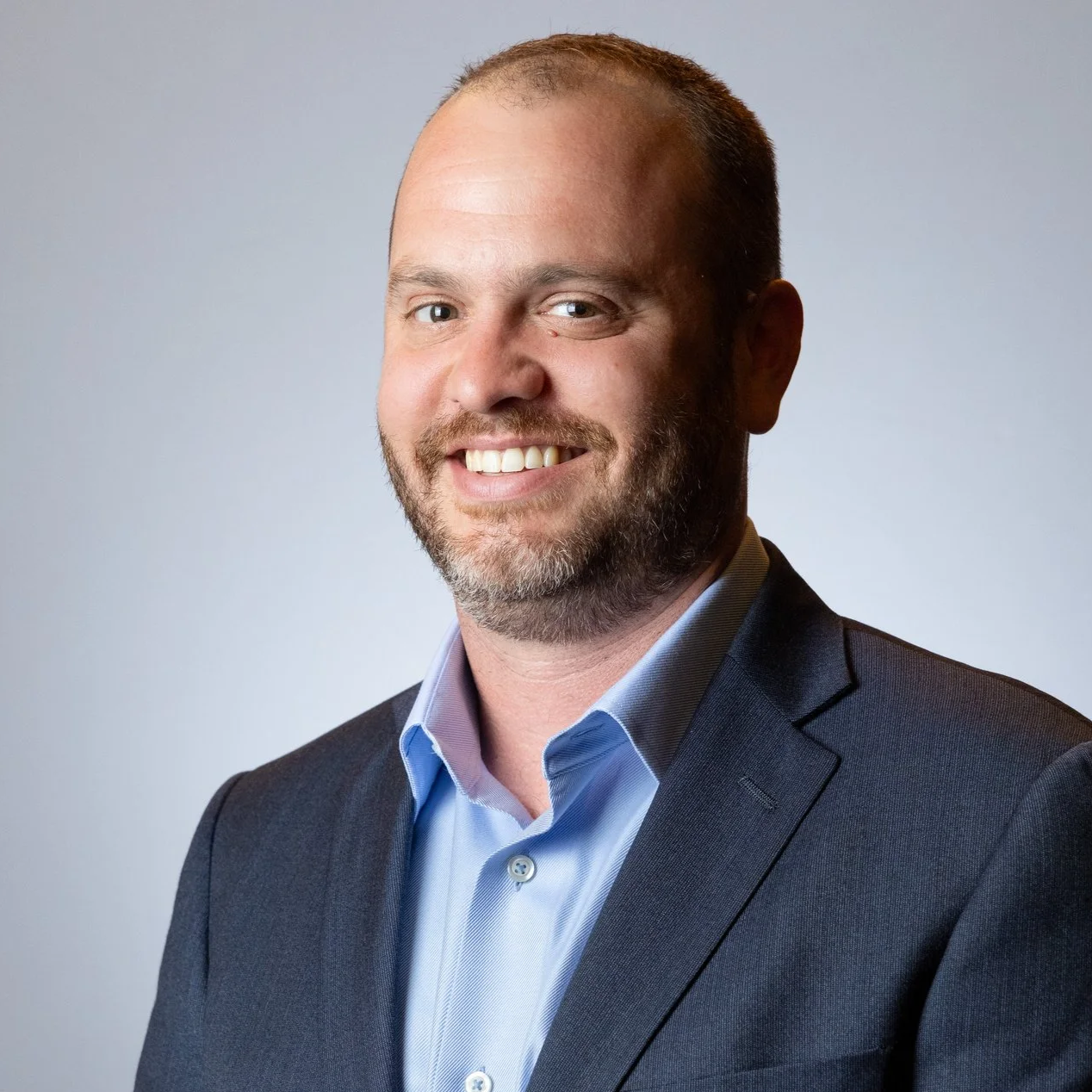 Noah Deich - senior advisor - with a beard and short hair, wearing a dark suit and light blue shirt, smiling against a plain gray background.
