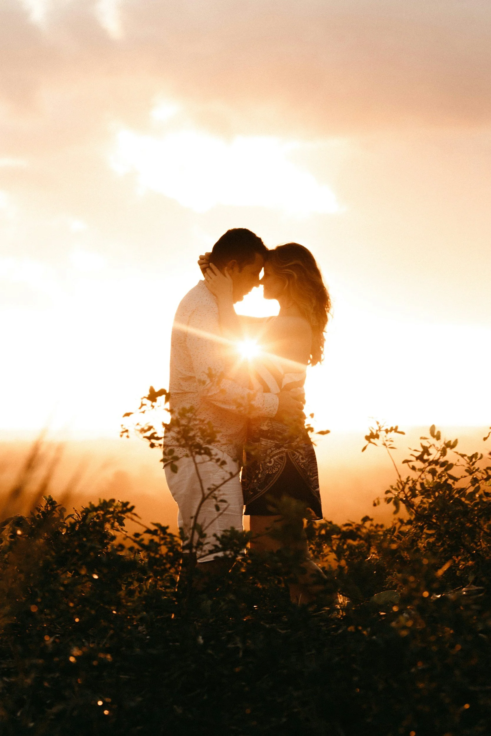 A couple is standing close together outdoors during sunset, touching foreheads, with the sun shining between them and their silhouettes visible against the sky.