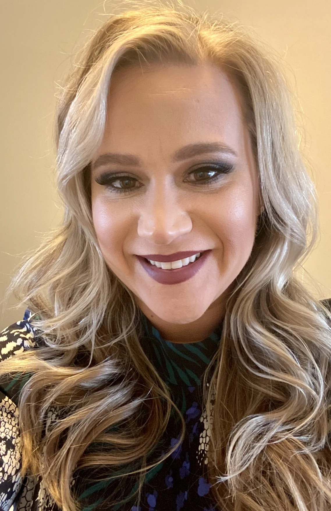 A woman with long, wavy blonde hair smiling toward the camera, wearing makeup and a dark patterned top.