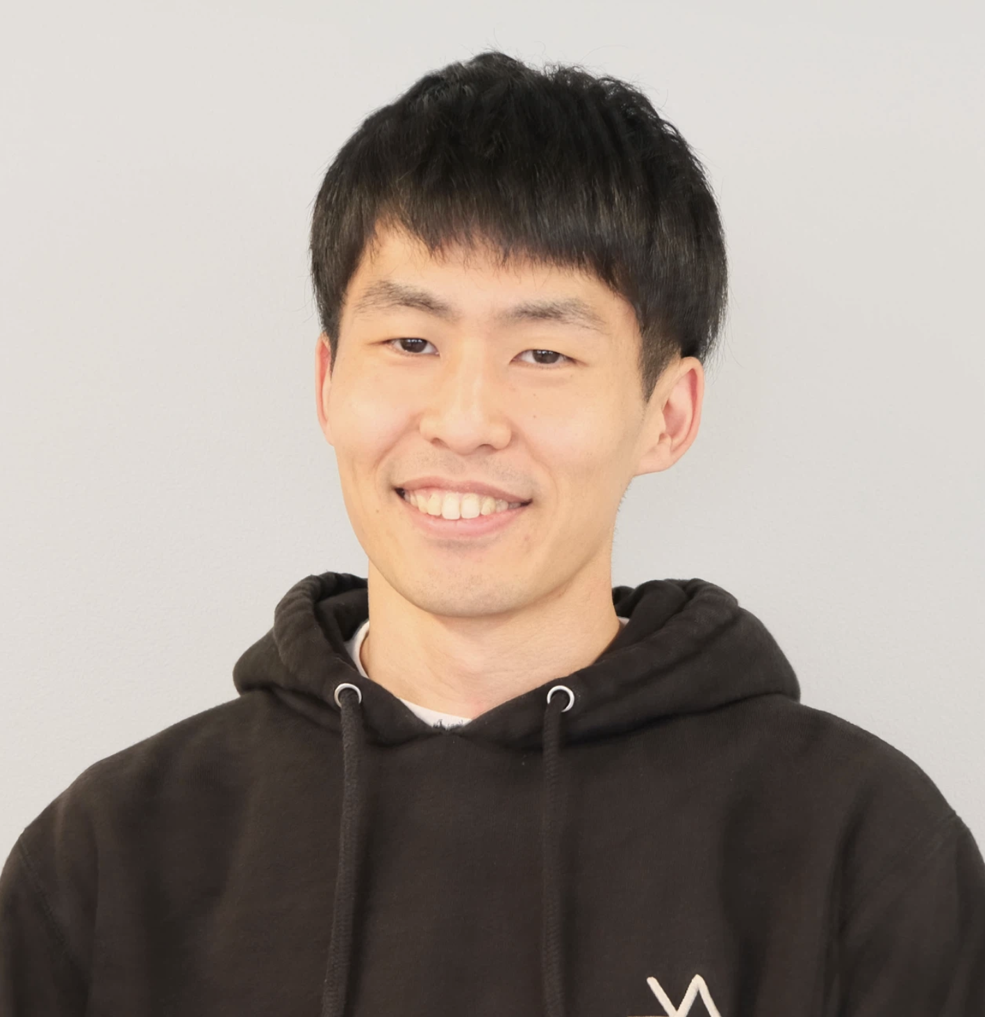 黒いパーカーを着た若い男性が笑顔でポーズを取っている。背景は白い壁。