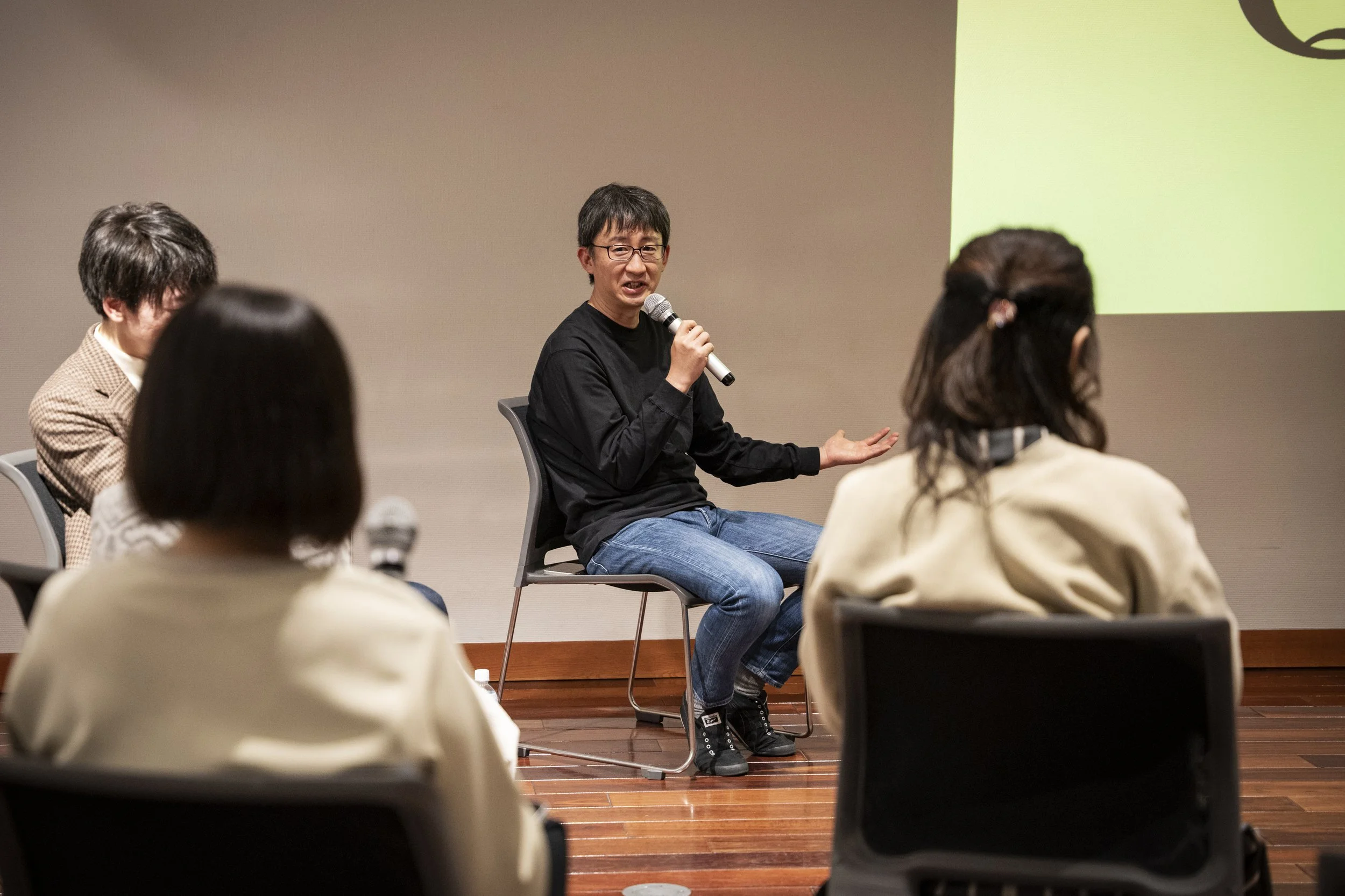 黒いシャツを着た男性がマイクを持ち、話している。数人の聴衆が座っている。