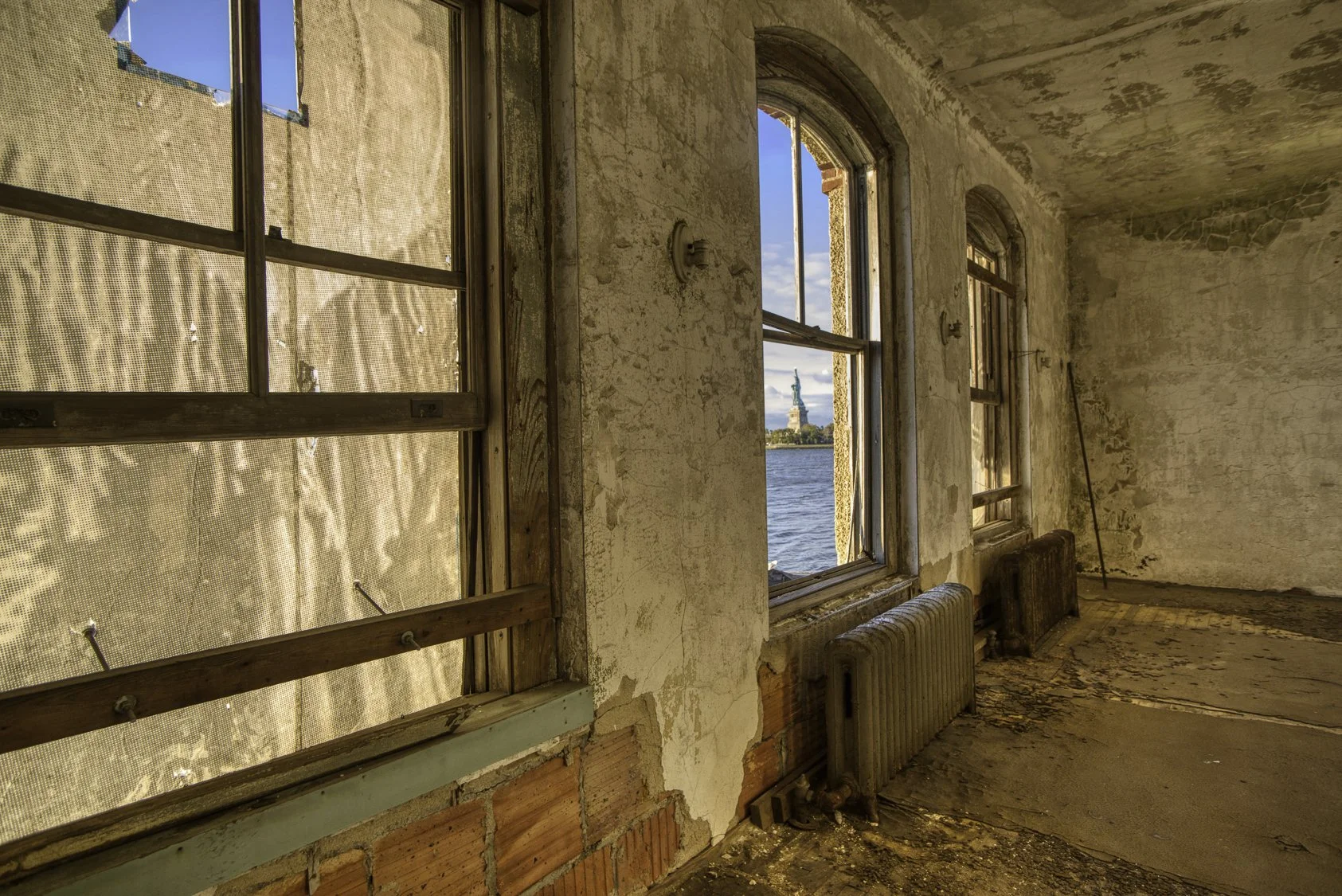 An interior view of an old, abandoned building with peeling paint on the walls, exposed brickwork, and large windows. Through the open window, the Statue of Liberty is visible across the water on a sunny day.