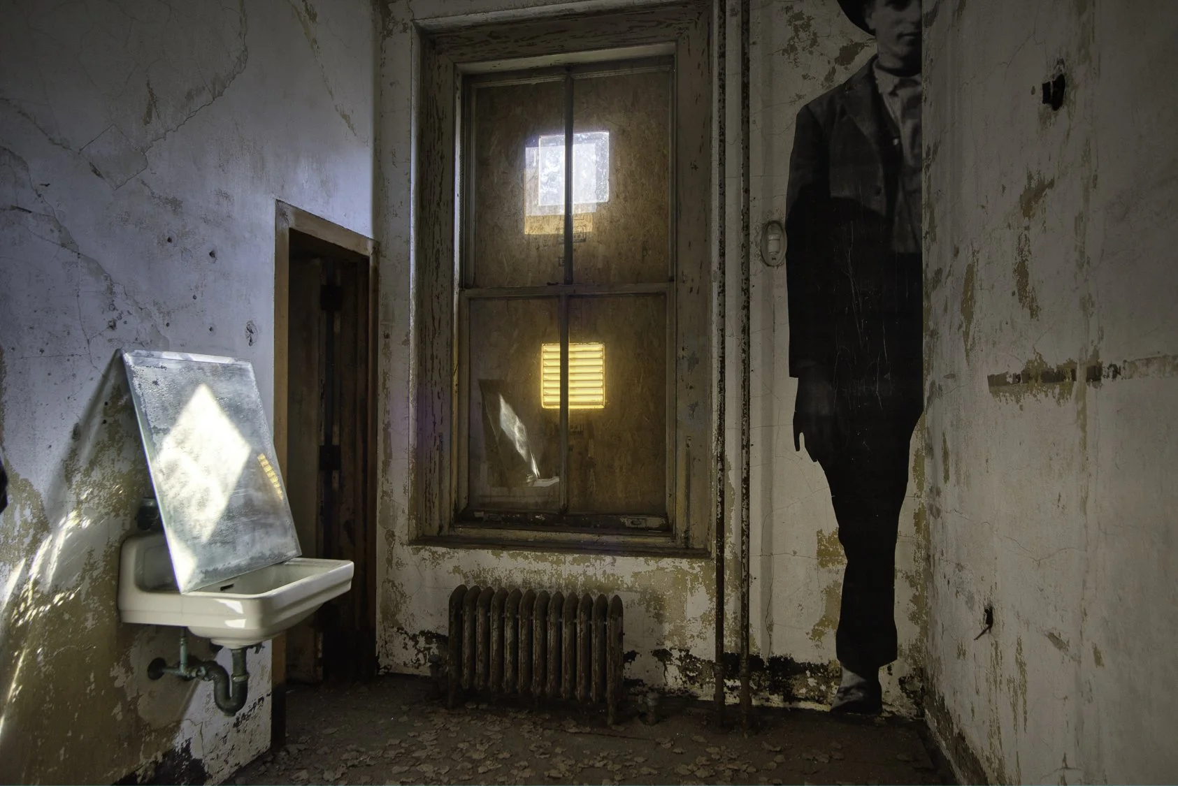 An abandoned room with peeling paint walls, a rusty radiator, a small sink with a mirror, a boarded-up window, and a large black and white poster of a man in formal attire on the wall.