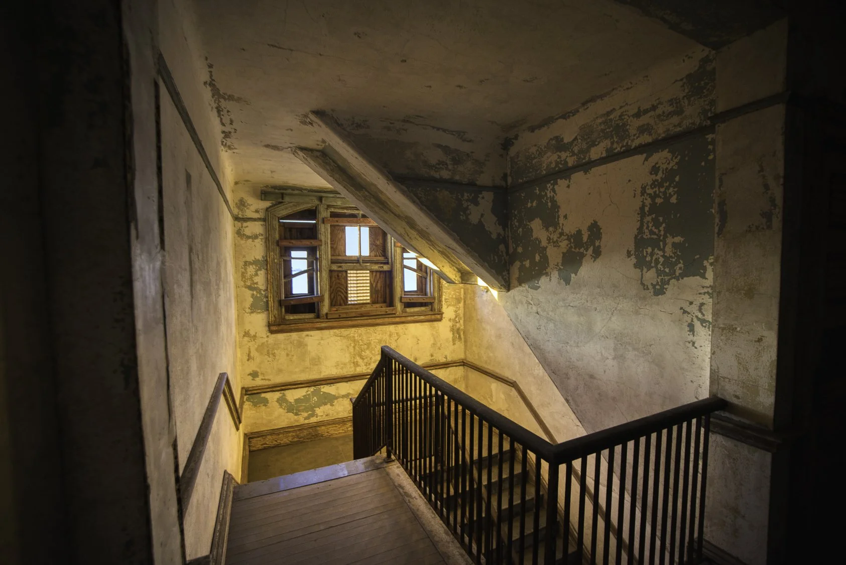 Old, worn staircase with peeling paint on walls, a window with shutters at the top of the stairs, and sunlight coming through the window.