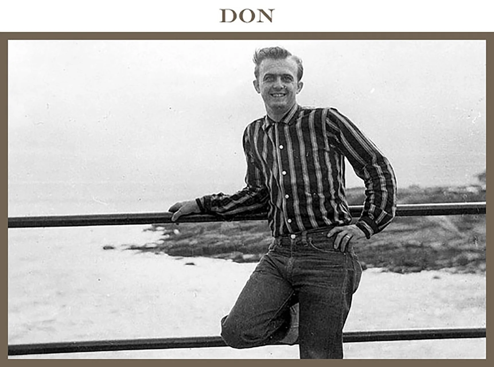 Black and white photo of a man standing outdoors by a railing, smiling at the camera, with a body of water and greenery in the background.